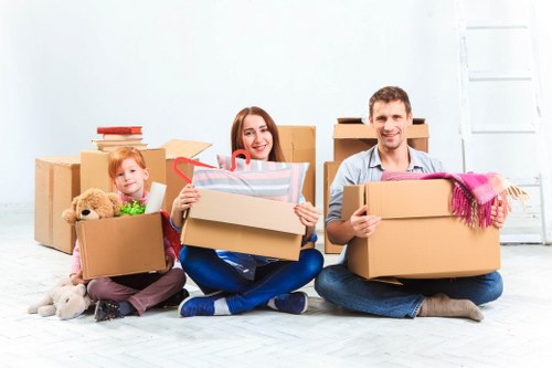 A family of three, comprising a young girl, a woman, and a man, are seated on the floor inside a bright, spacious room with white walls, surrounded by numerous cardboard moving boxes in various sizes. The young girl, positioned on the left, has red hair and is holding a box filled with children's toys, including a teddy bear and a green stuffed animal, with some toys spilling out. The woman in the centre is smiling, holding a medium-sized box with a transparent bag of miscellaneous household items inside, and she is dressed in a grey top and blue jeans. The man on the right, also smiling, is holding a large box wrapped with a pink scarf draped over his arm, dressed in a light-colored shirt and dark jeans. Behind them, some boxes are stacked on the floor, and others are placed on what appears to be a simple, uncluttered interior space suitable for a house move. The scene suggests an active packing or unpacking process, consistent with house removals services in the London area. The room is well-lit with natural light, and the setting emphasizes preparation for a residential relocation within Kent and nearby areas.