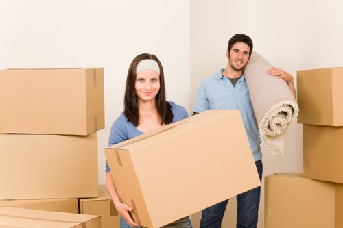 A young woman and a young man, both with light skin, are standing inside a residential space, surrounded by stacked cardboard boxes in various sizes, suggesting a house moving or removals scenario. The woman is holding a medium-sized rectangular cardboard box, smiling at the camera. She has straight, shoulder-length dark hair and wears a light blue shirt under a darker cardigan. The man, standing slightly behind her on the right, is carrying a rolled-up beige carpet with a textured surface over his shoulder, also smiling. He has short, dark hair and is dressed in a light blue shirt and dark trousers. The background features a plain, off-white wall and a minimalistic interior setting, indicative of a clean and organized home environment in Kentish Town, London. The scene is well-lit with natural or soft lighting, emphasizing the professional and neutral nature of the household move, characteristic of Kentish Town Man with Van services. The overall image captures the process of packing and preparing household items for transport, reflecting the company's specialized removals services within the London Borough of Camden.