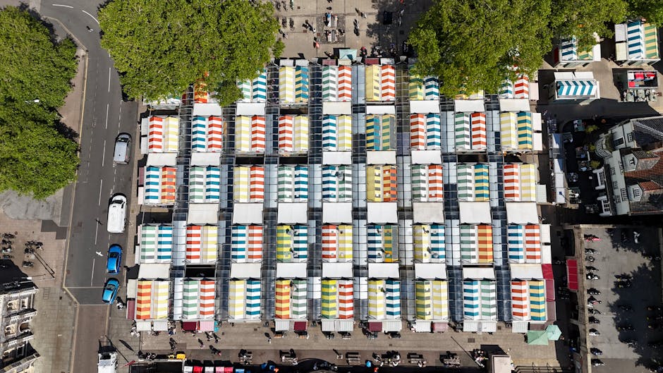 An aerial view of a large outdoor market with numerous tightly packed stalls arranged in a grid pattern, each stall covered with colorful striped awnings in shades of red, blue, yellow, and green. The